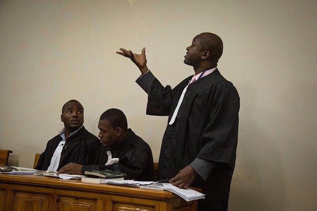 A lawyer wearing a black robe and a pink business shirt standing up in court addressing the room.